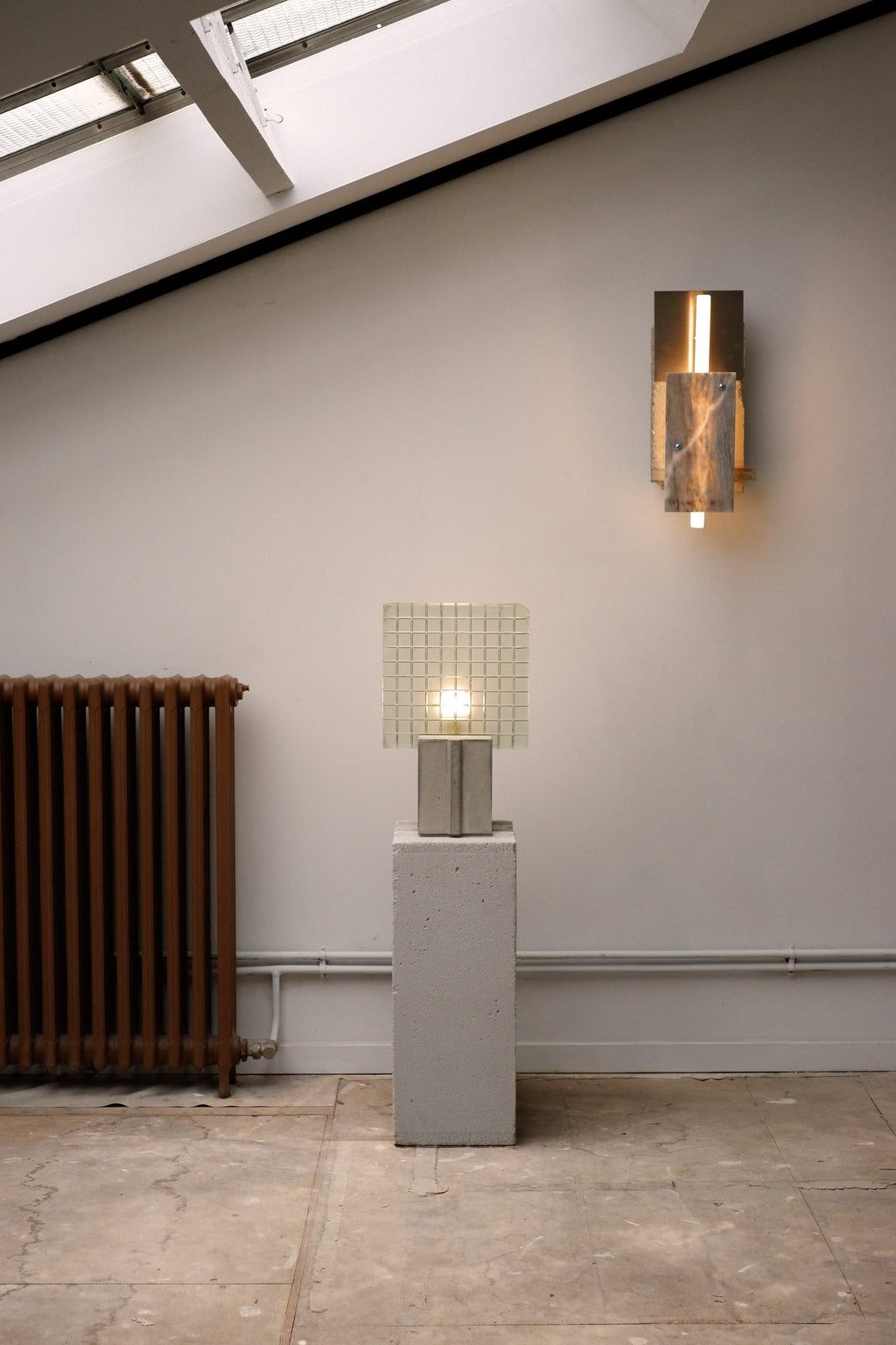 A floor lamp with a concrete base, glass shade, and wood details, standing next to a wall-mounted lamp with a similar design.