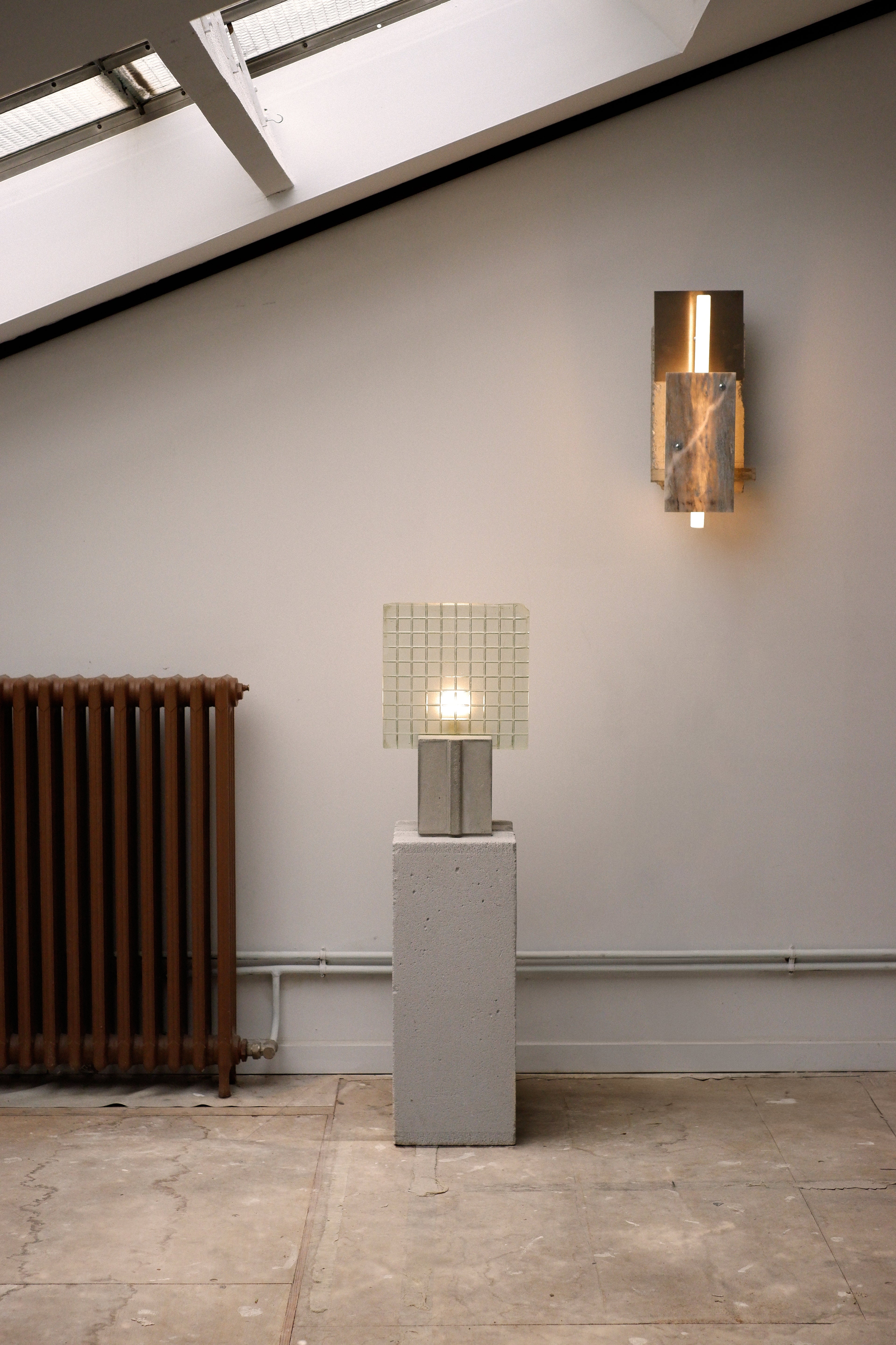 A floor lamp with a concrete base, glass shade, and wood details, standing next to a wall-mounted lamp with a similar design.